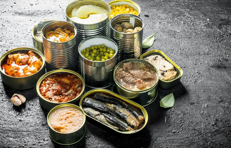 Auf einer Schieferplatte stehen unterschiedliche geöffnete Konservendosen, unter anderem mit Ananas, Pilzen, Gemüse und Fisch.