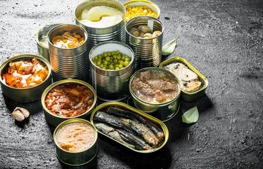 Auf einer Schieferplatte stehen unterschiedliche geöffnete Konservendosen, unter anderem mit Ananas, Pilzen, Gemüse und Fisch.