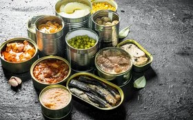 Auf einer Schieferplatte stehen unterschiedliche geöffnete Konservendosen, unter anderem mit Ananas, Pilzen, Gemüse und Fisch.