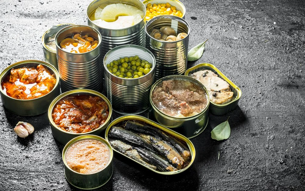 Auf einer Schieferplatte stehen unterschiedliche geöffnete Konservendosen, unter anderem mit Ananas, Pilzen, Gemüse und Fisch.