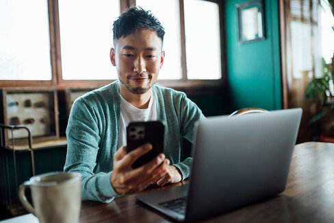 Ein junger Mann sitzt am Schreibtisch. Via Laptop und Smartphone sucht er online nach einem Arzt in seiner Nähe.