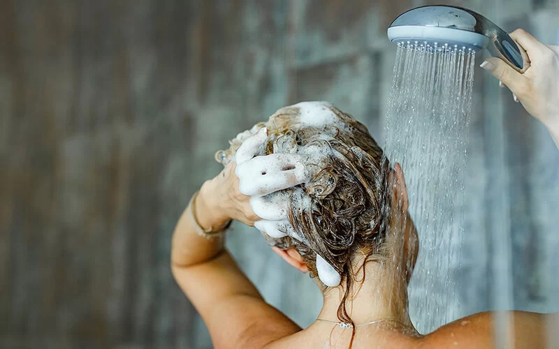 Eine Frau steht unter der Dusche, zu sehen ist ihr Hinterkopf. Ihre Haare sind eingeschäumt. Mit der Hand fährt sie sich durch die Haare.