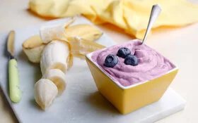 Schneidebrett auf dem eine halbgeschälte und geschnittene Banane liegt. Daneben ein Schälchen mit Bananen-Blaubeer-Eiscreme.