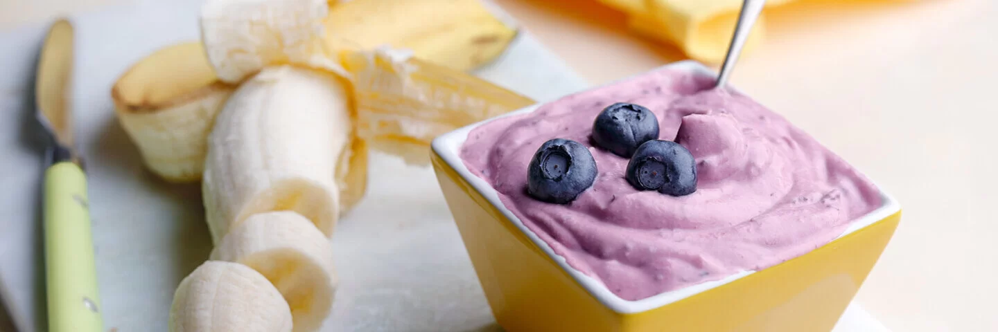 Schneidebrett auf dem eine halbgeschälte und geschnittene Banane liegt. Daneben ein Schälchen mit Bananen-Blaubeer-Eiscreme.