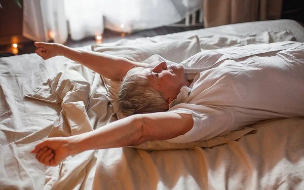 Eine Senorin in einem weißen Oberteil führt eine Streckübung im Bett durch.