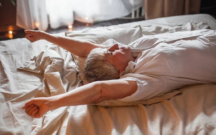 Eine Senorin in einem weißen Oberteil führt eine Streckübung im Bett durch.