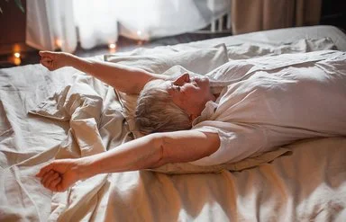 Eine Senorin in einem weißen Oberteil führt eine Streckübung im Bett durch.