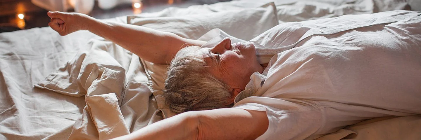 Eine Senorin in einem weißen Oberteil führt eine Streckübung im Bett durch.