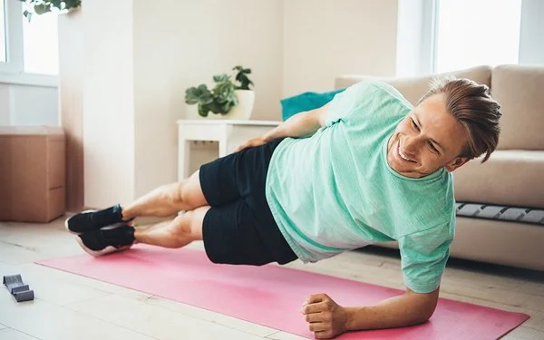 Ein lächelnder Mann hält einen Seitstütz auf einer Yogamatte in einem Wohnzimmer.