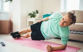 Ein lächelnder Mann hält einen Seitstütz auf einer Yogamatte in einem Wohnzimmer.