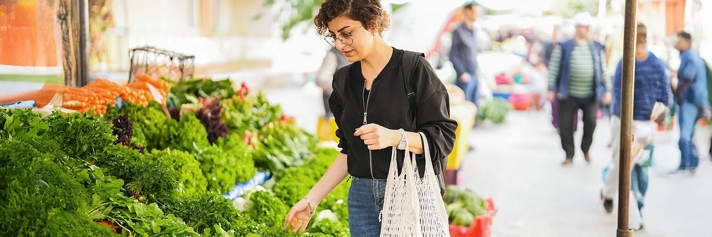 Eine Frau steht vor einem Gemüsestand auf dem Wochenmarkt und betrachtet die ausliegenden Blattsalate. An ihrem linken Arm hängen zwei Einkaufsnetze mit Obst.