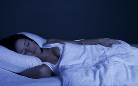 Einschlafhilfe: Eine junge Frau schläft entspannt in einem abgedunkelten Raum mit einem stützenden Kissen unter dem Kopf. 