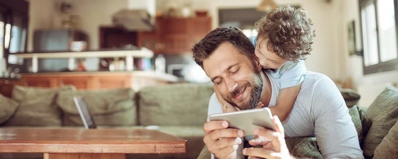 Papa und Sohn liegen auf dem Sofa. Papa hält ein Handy in der Hand. Der Sohn umarmt seinen Vater.