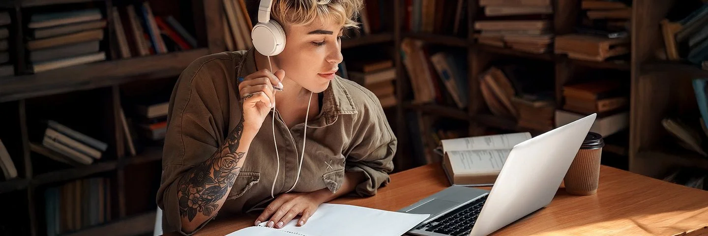 Eine junge Frau mit Kopfhörern sitzt vor einem Laptop und macht Notizen.