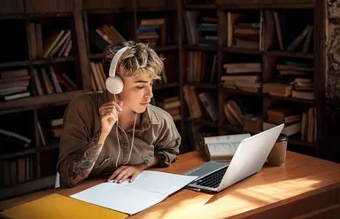 Eine junge Frau mit Kopfhörern sitzt vor einem Laptop und macht Notizen.