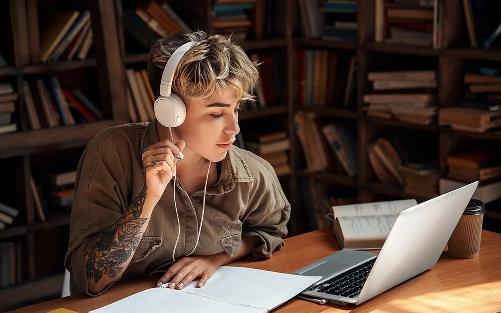 Eine junge Frau mit Kopfhörern sitzt vor einem Laptop und macht Notizen.