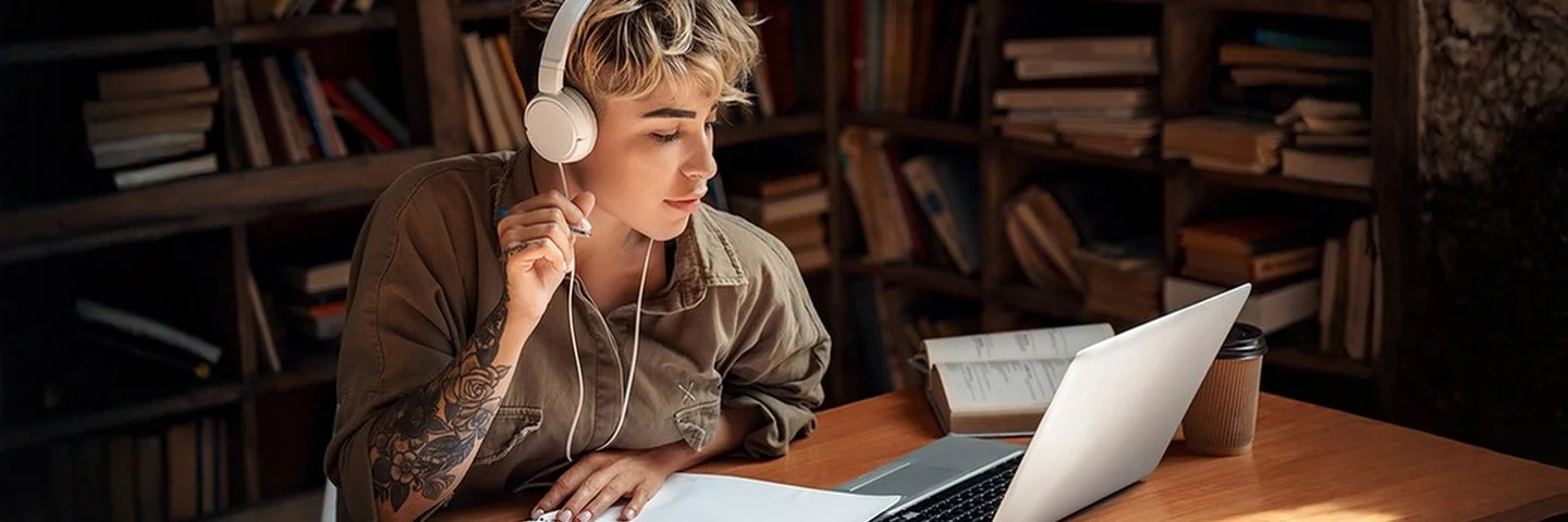 Eine junge Frau mit Kopfhörern sitzt vor einem Laptop und macht Notizen.