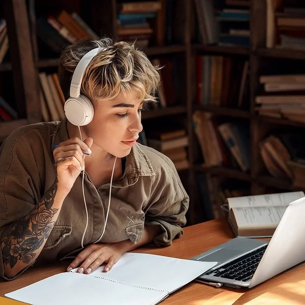 Eine junge Frau mit Kopfhörern sitzt vor einem Laptop und macht Notizen.
