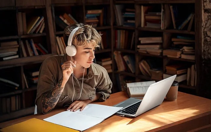 Eine junge Frau mit Kopfhörern sitzt vor einem Laptop und macht Notizen.