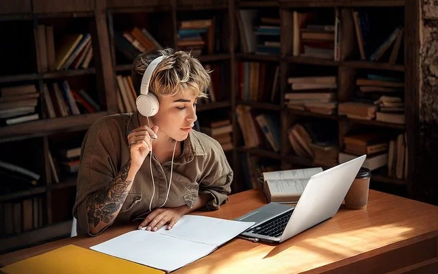 Eine junge Frau mit Kopfhörern sitzt vor einem Laptop und macht Notizen.