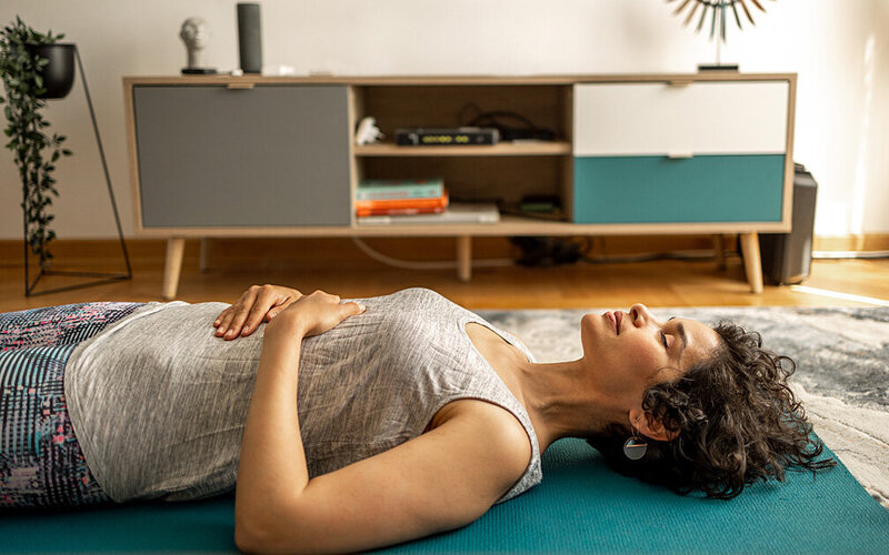 Frau liegt mit geschlossenen Augen auf einer Yogamatte und ihre Hände liegen auf dem Bauch.
