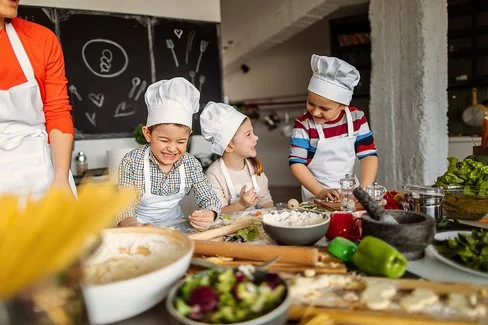 Drei fröhliche Kinder in Kochmützen und Schürzen kochen gemeinsam in einer Schulküche. Neben ihnen steht eine Lehrerin.