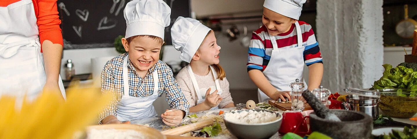 Drei fröhliche Kinder in Kochmützen und Schürzen kochen gemeinsam in einer Schulküche. Neben ihnen steht eine Lehrerin.