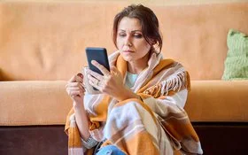 Eine Frau in einem Wohnzimmer. Sie ist in eine Wolldecke eingehüllt. In der rechten Hand hält sie eine Tasse, aus der ein Teebeutel heraushängt, in der linken Hand ein Handy, auf dem sie aufmerksam liest.