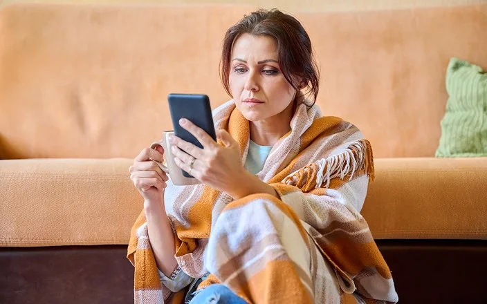 Eine Frau in einem Wohnzimmer. Sie ist in eine Wolldecke eingehüllt. In der rechten Hand hält sie eine Tasse, aus der ein Teebeutel heraushängt, in der linken Hand ein Handy, auf dem sie aufmerksam liest.