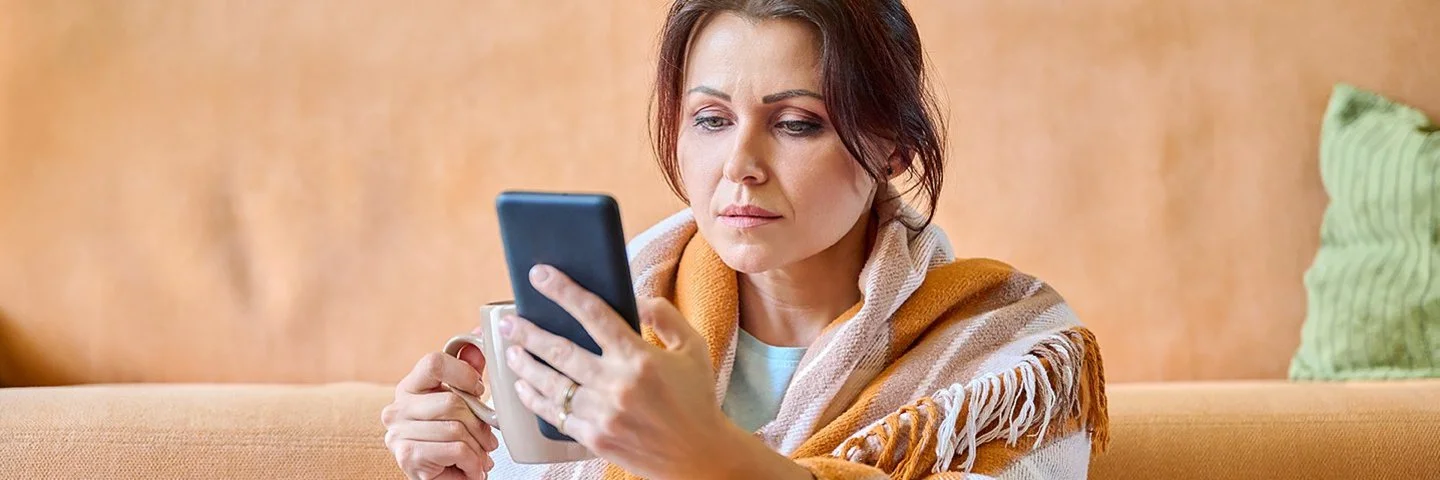 Eine Frau in einem Wohnzimmer. Sie ist in eine Wolldecke eingehüllt. In der rechten Hand hält sie eine Tasse, aus der ein Teebeutel heraushängt, in der linken Hand ein Handy, auf dem sie aufmerksam liest.