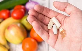 In einer Handfläche liegen verschiedene Tabletten, Pillen und Dragees, im Hintergrund sind verschwommen frisches Obst und Gemüse zu erkennen.