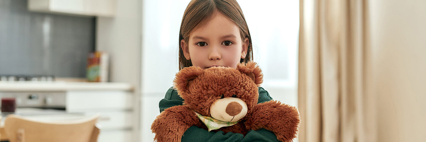 Ein kleines Mädchen drückt seinen Teddybären fest an sich und schaut schüchtern nach vorne.