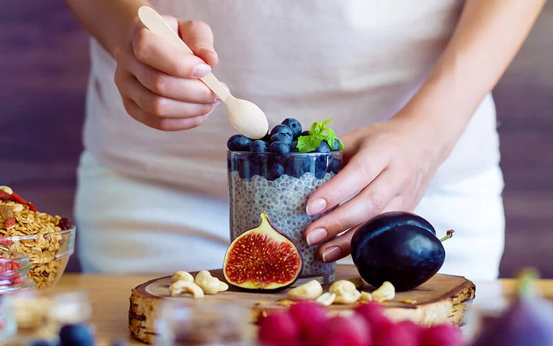 Eine Person bereitet ein gesundes Frühstück mit Joghurt, Chia und Blaubeeren. Auf dem Tisch liegen weitere Zutaten wie Sommerbeeren, Obst und Nüsse.