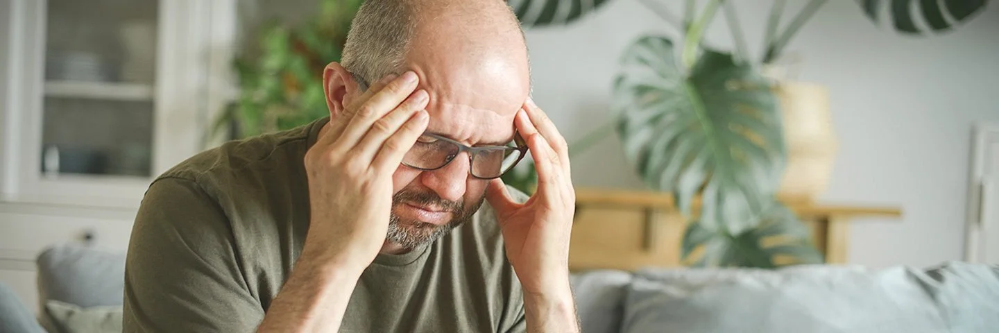 Ein Mann mittleren Alters sitzt leicht nach vorne gebeugt zu Hause auf der Couch und fasst sich mit den Händen an den Kopf.