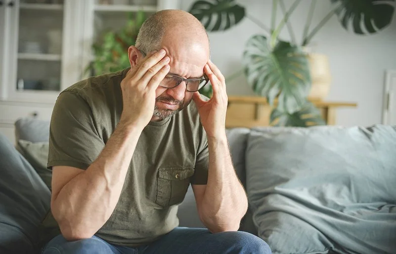 Ein Mann mittleren Alters sitzt leicht nach vorne gebeugt zu Hause auf der Couch und fasst sich mit den Händen an den Kopf.