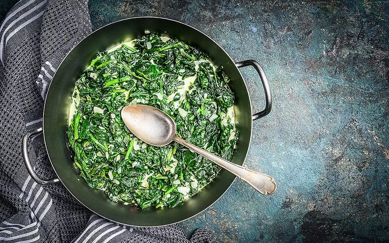 Pfanne mit gedünstetem Rahmspinat und einem Löffel auf einer dunklen Arbeitsfläche
