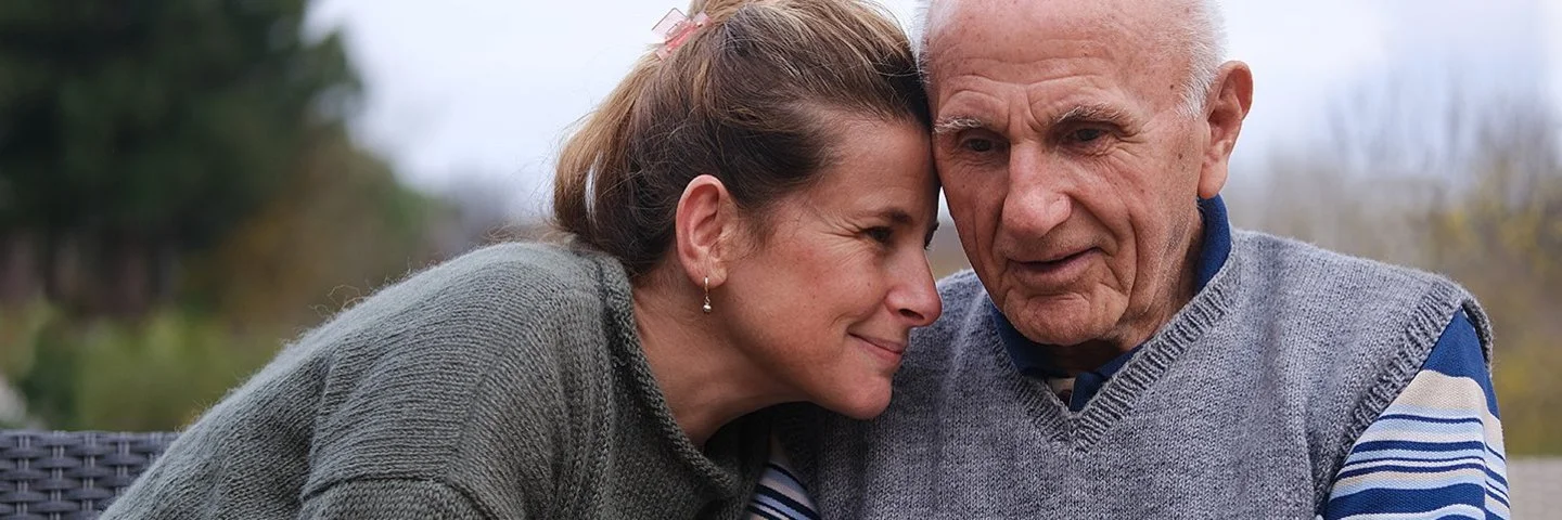 Eine Frau sitzt ganz nah neben ihrem alten Vater. Sie hält seine Hände und hat ihre Stirn an seine Stirn gelegt. Sie befinden sich draußen. Im Hintergrund ist eine kühle Herbststimmung zu sehen.