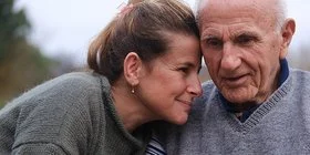 Eine Frau sitzt ganz nah neben ihrem alten Vater. Sie hält seine Hände und hat ihre Stirn an seine Stirn gelegt. Sie befinden sich draußen. Im Hintergrund ist eine kühle Herbststimmung zu sehen.