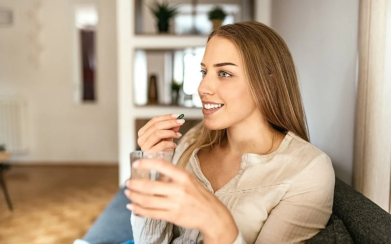 Eine junge Frau mit langen braunen Haaren sitzt in einem Wohnzimmer auf dem Sofa und hält in einer Hand eine Hartkapsel, in der anderen ein Glas Wasser.