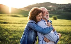 Ein älteres lachendes Paar umarmt sich vor einer hügeligen Kulisse bei tiefstehender Sonne.