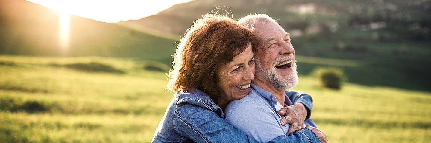Ein älteres lachendes Paar umarmt sich vor einer hügeligen Kulisse bei tiefstehender Sonne.