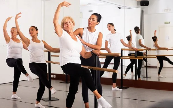 Eine Gruppe von Frauen tanzt Ballett. Eine ältere Frau bekommt dabei eine Einleitung durch eine Lehrerin.