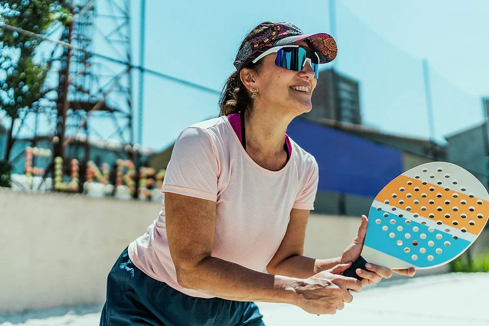 An einem sonnigen Tag steht eine Frau mit einem Beachballschläger in den Händen auf einem Sportplatz. Sie trägt ein Stirnband mit Sonnenschild, eine Sonnenbrille und ein T-Shirt.