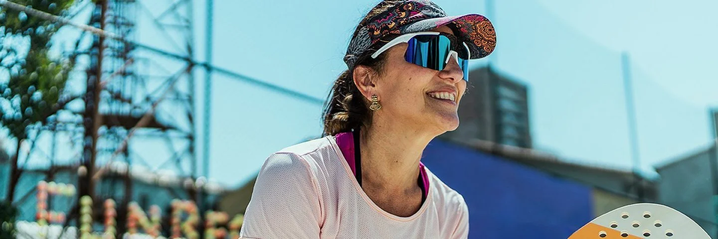 An einem sonnigen Tag steht eine Frau mit einem Beachballschläger in den Händen auf einem Sportplatz. Sie trägt ein Stirnband mit Sonnenschild, eine Sonnenbrille und ein T-Shirt.