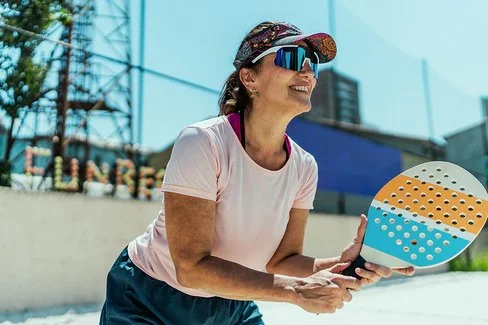An einem sonnigen Tag steht eine Frau mit einem Beachballschläger in den Händen auf einem Sportplatz. Sie trägt ein Stirnband mit Sonnenschild, eine Sonnenbrille und ein T-Shirt.
