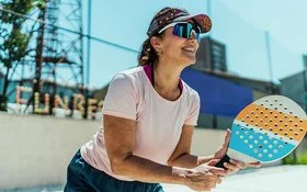 An einem sonnigen Tag steht eine Frau mit einem Beachballschläger in den Händen auf einem Sportplatz. Sie trägt ein Stirnband mit Sonnenschild, eine Sonnenbrille und ein T-Shirt.