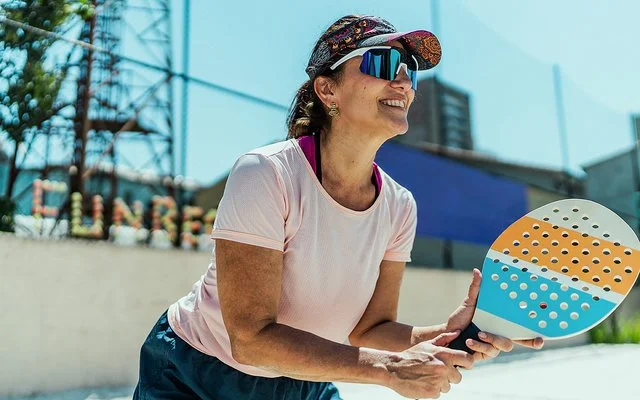 An einem sonnigen Tag steht eine Frau mit einem Beachballschläger in den Händen auf einem Sportplatz. Sie trägt ein Stirnband mit Sonnenschild, eine Sonnenbrille und ein T-Shirt.