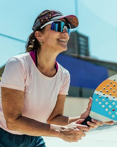 An einem sonnigen Tag steht eine Frau mit einem Beachballschläger in den Händen auf einem Sportplatz. Sie trägt ein Stirnband mit Sonnenschild, eine Sonnenbrille und ein T-Shirt.