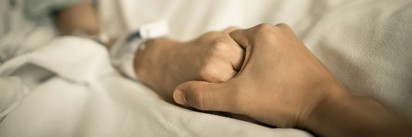 Eine Person hält die Hand einer anderen Person, die in einem Krankenhausbett liegt.