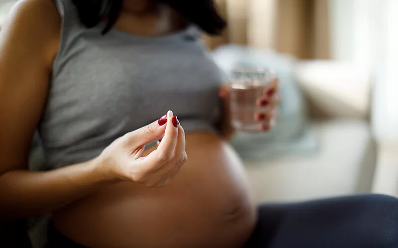 Eine schwangere Frau sitzt mit kurzem Oberteil auf dem Sofa und hält eine Tablette sowie ein Glas Wasser in den Händen.